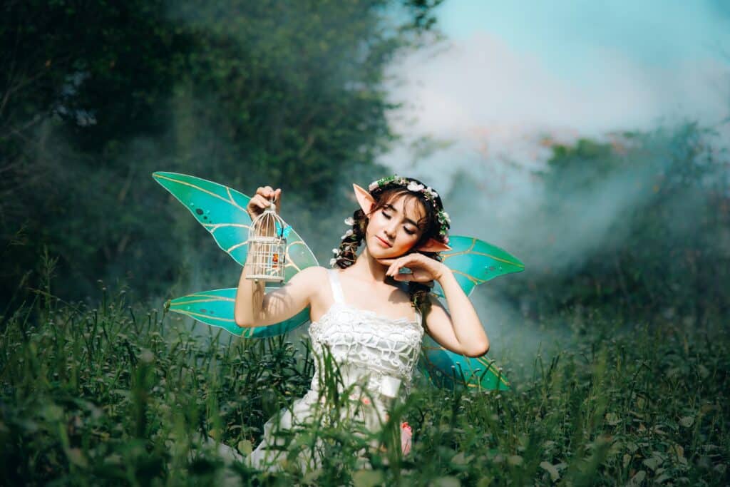 A person dressed as a fairy with blue wings and elf ears, holding a small cage, poses in a misty green field.