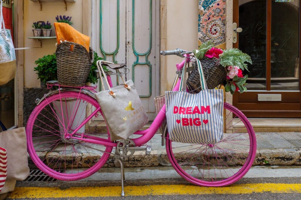 A pink bicycle with baskets carrying flowers and bags, including one that says "Dream Big," is parked on a street near a decorated building entrance—an inspiring symbol of ideas for sustainable living.