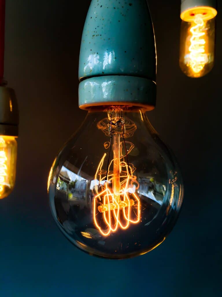 Close-up of a glowing incandescent light bulb with a visible filament against a dark background, demonstrating ways to conserve energy beside two similar bulbs.