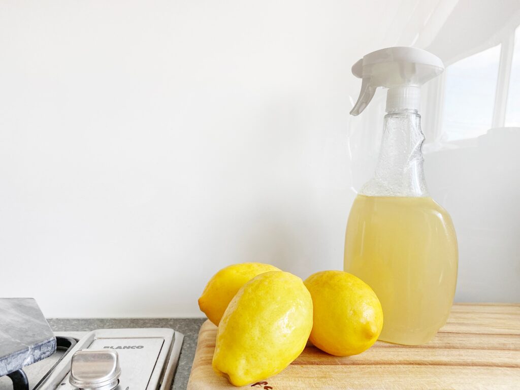 A spray bottle with lemon juice and three whole lemons sits on a wooden cutting board, ready to tackle your summer cleaning checklist.