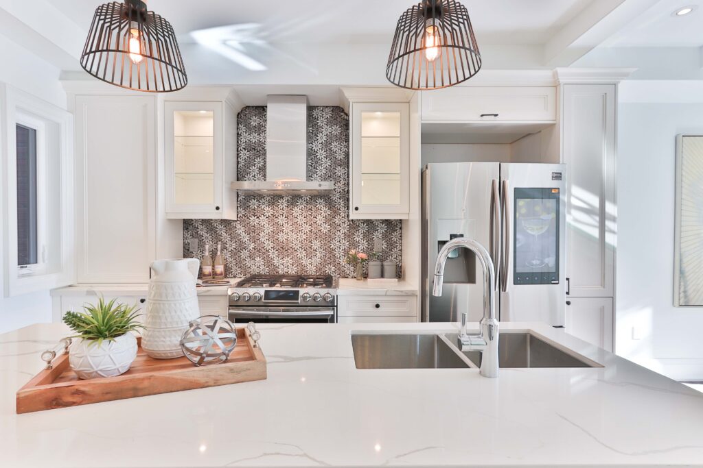 Modern kitchen with white cabinets, a stainless steel fridge, and a double sink. Decorative backsplash behind the stove and pendant lights overhead. Countertop with a tray of decor items.
