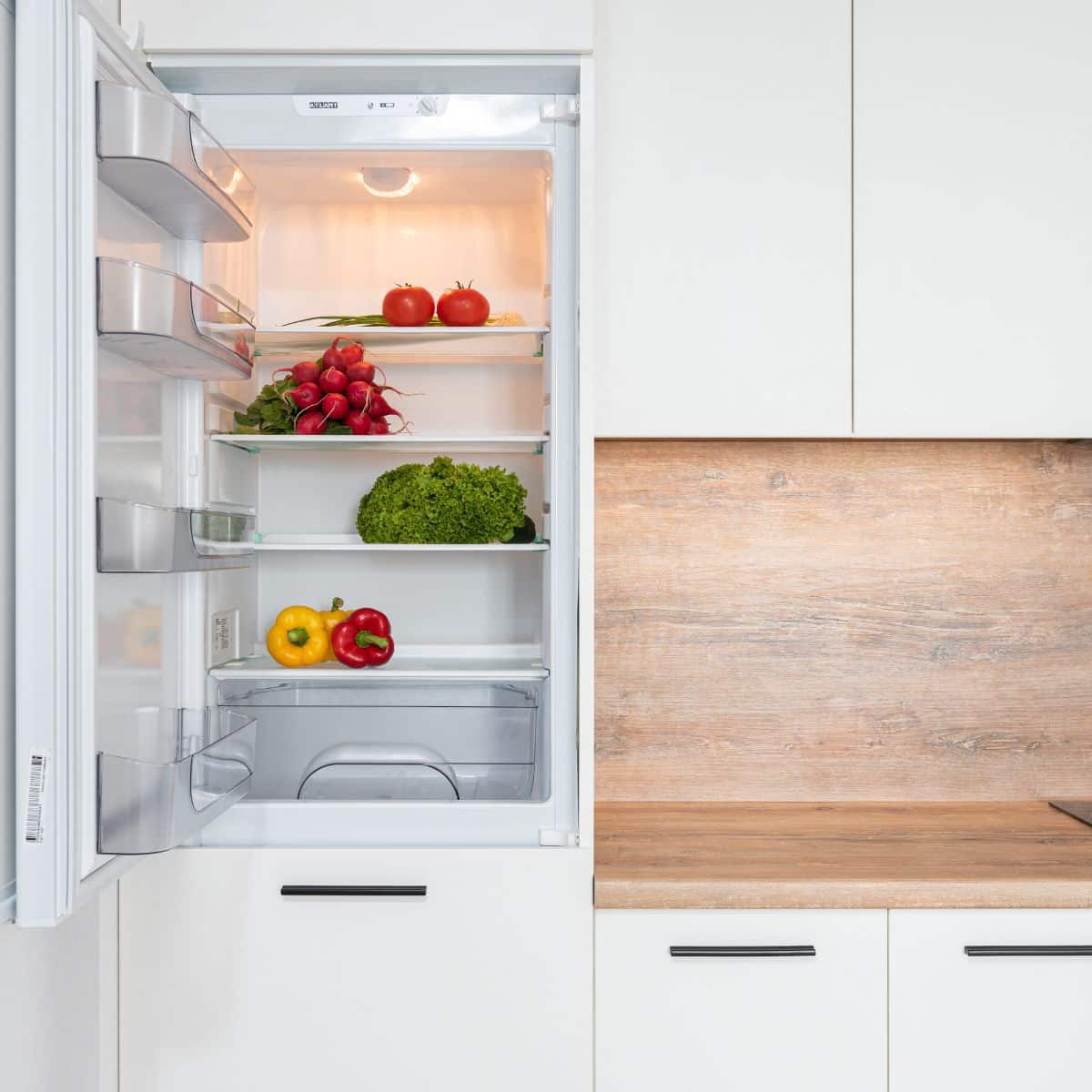 Open fridge revealing fresh vegetables like tomatoes, radishes, lettuce, and peppers. Beside the crisp produce, white kitchen cabinets and a wooden backsplash add a touch of style. Perfectly organized for those learning how to deep clean your fridge.