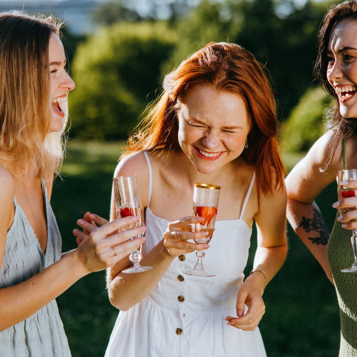 Three women laughing and holding glasses of drinks outdoors with greenery in the background.