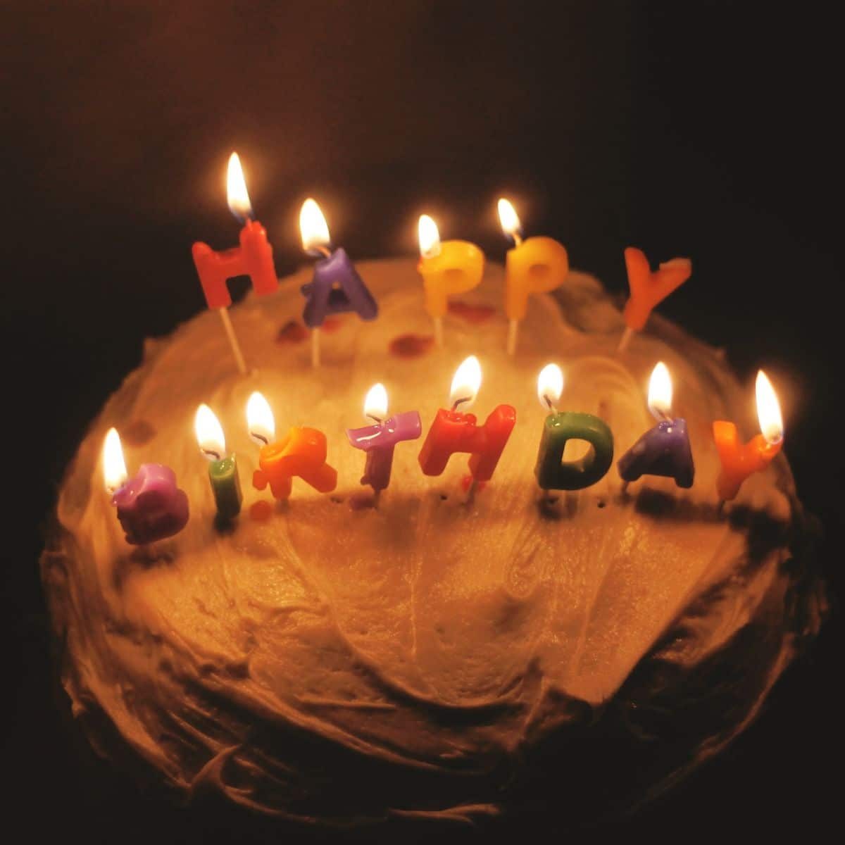 A cake with white frosting is topped with lit birthday candles spelling "HAPPY BIRTHDAY.