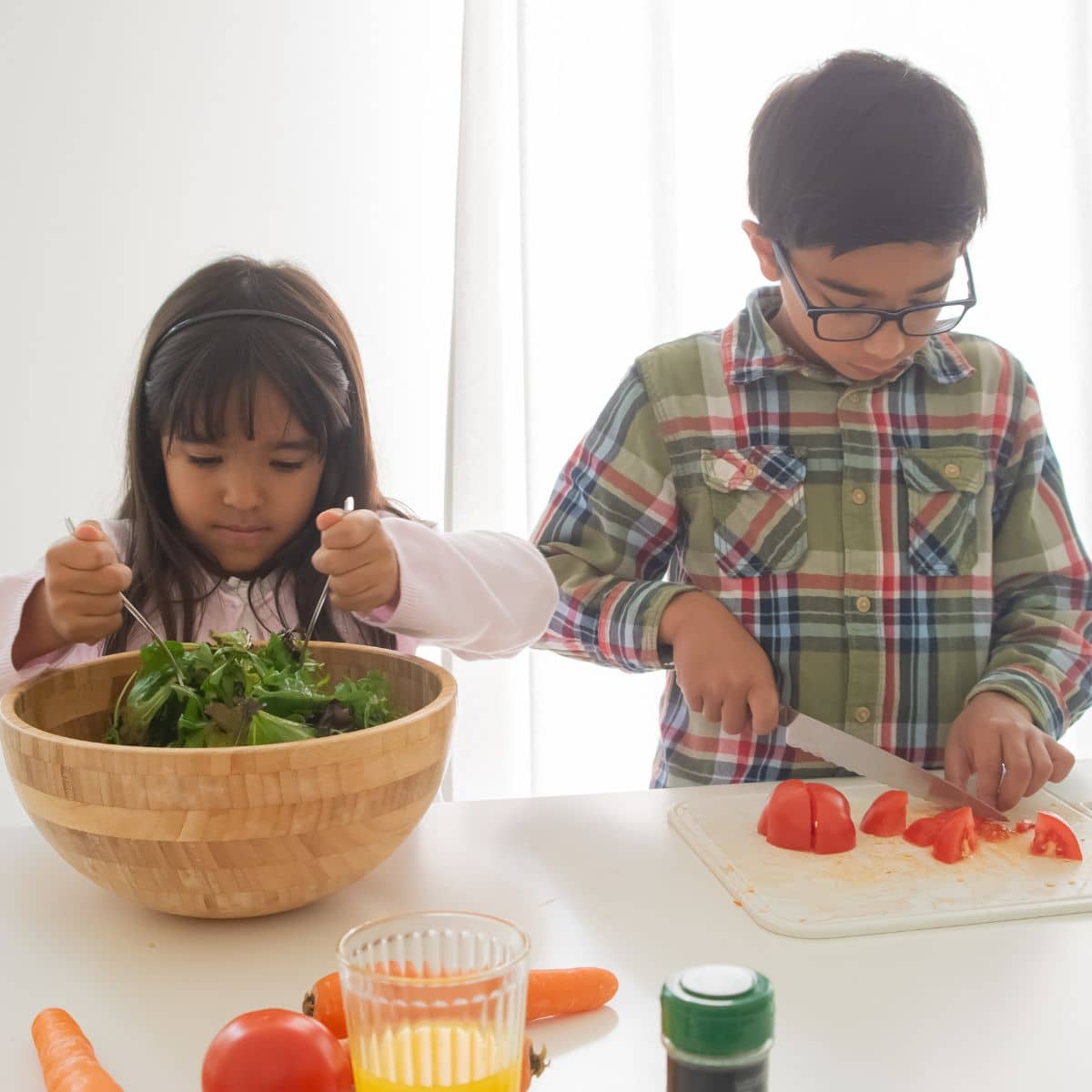 Two children prepare food in a bright kitchen. The girl mixes a salad in a wooden bowl, while the boy slices tomatoes on a cutting board. A glass and extra vegetables are on the table.
