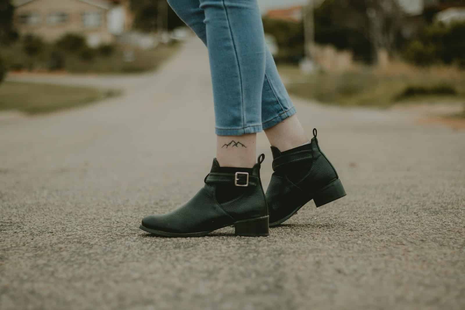 Person wearing black ankle boots and rolled-up jeans, standing on a paved road with a mountain tattoo visible above the boot.