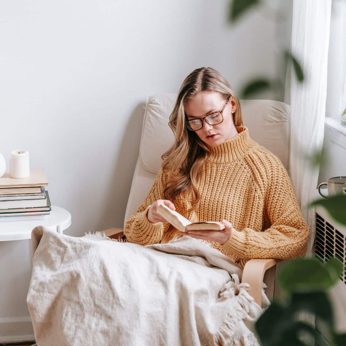 A person wearing glasses and a yellow sweater reads a book while sitting in a chair, wrapped in a blanket. Nearby, there is a small table with books and a candle.