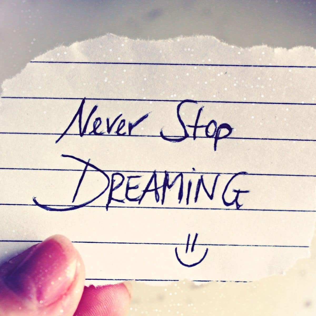 Hand holding a torn piece of lined paper with the handwritten message "Never Stop Dreaming" and a small smiley face.