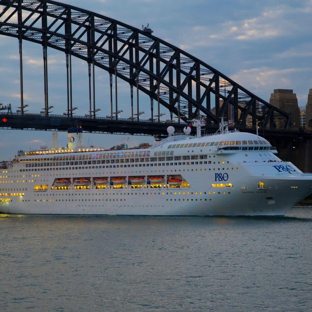 A large P&O cruise ship sails near a steel arch bridge at dusk, with city buildings in the background.