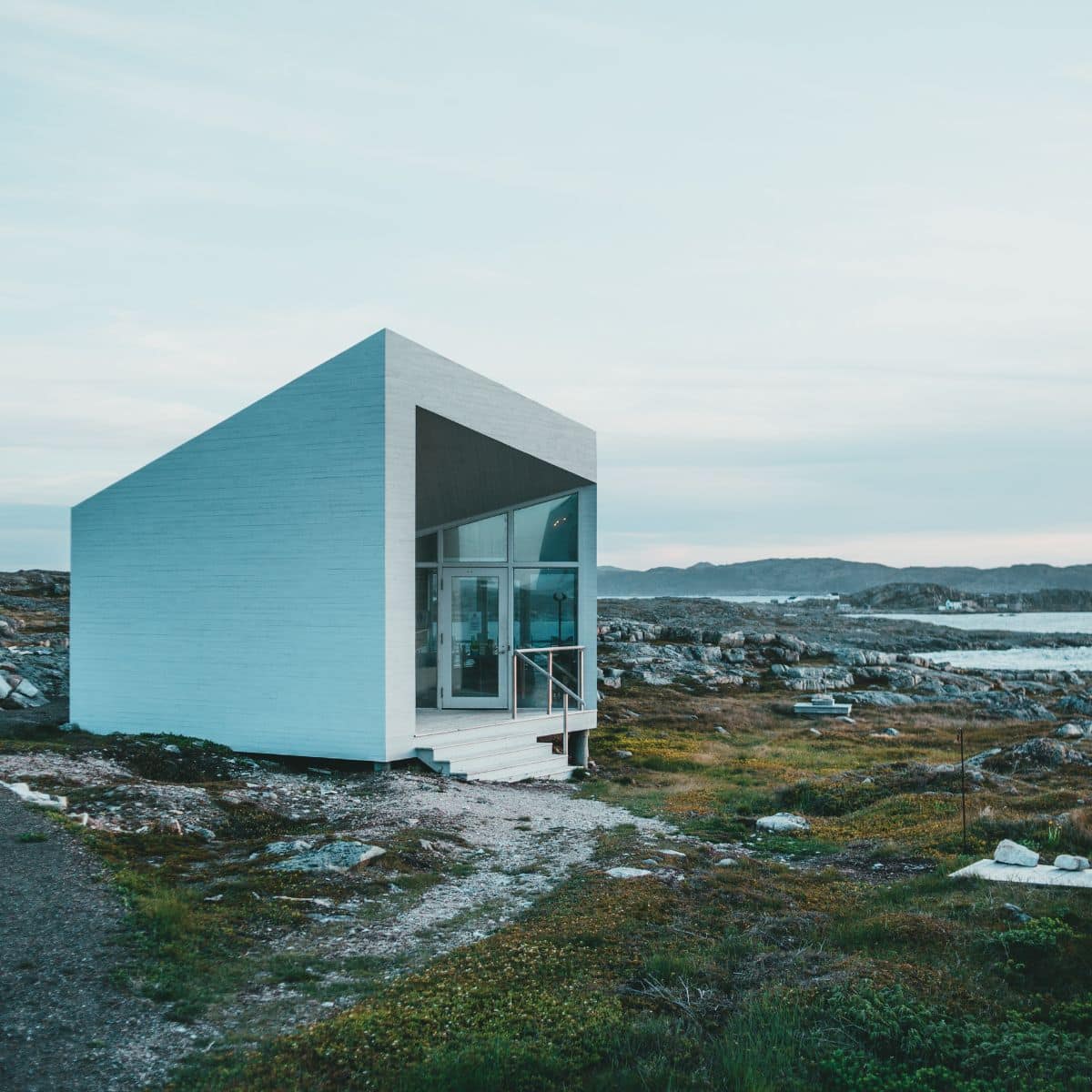 Modern white geometric building with large glass windows situated in a rocky coastal landscape under a pale blue sky.