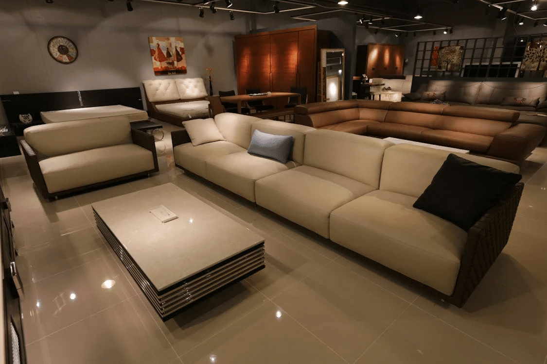 A modern furniture showroom with beige and brown sofas, a coffee table, and artwork on the walls, illuminated by spotlights.