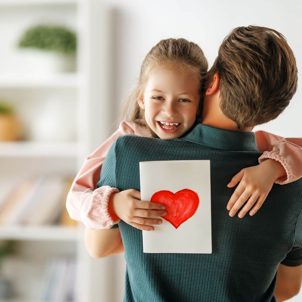 A smiling girl hugs a man while holding a card with a red heart.