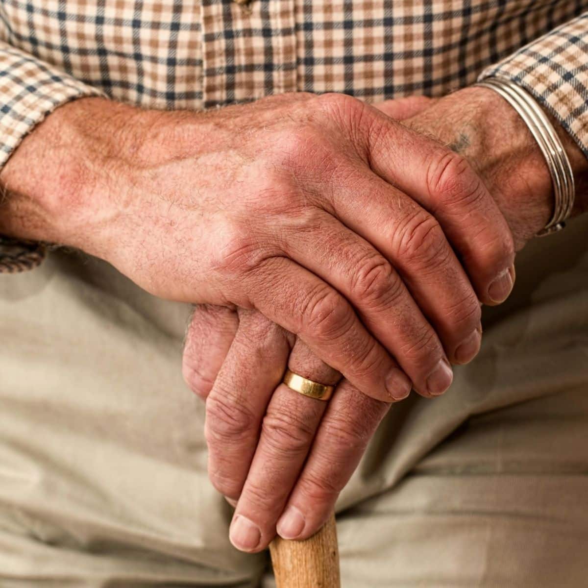 Close-up of an older person's hands resting on a wooden cane, wearing a gold ring and a metal bracelet, dressed in a plaid shirt and light pants.