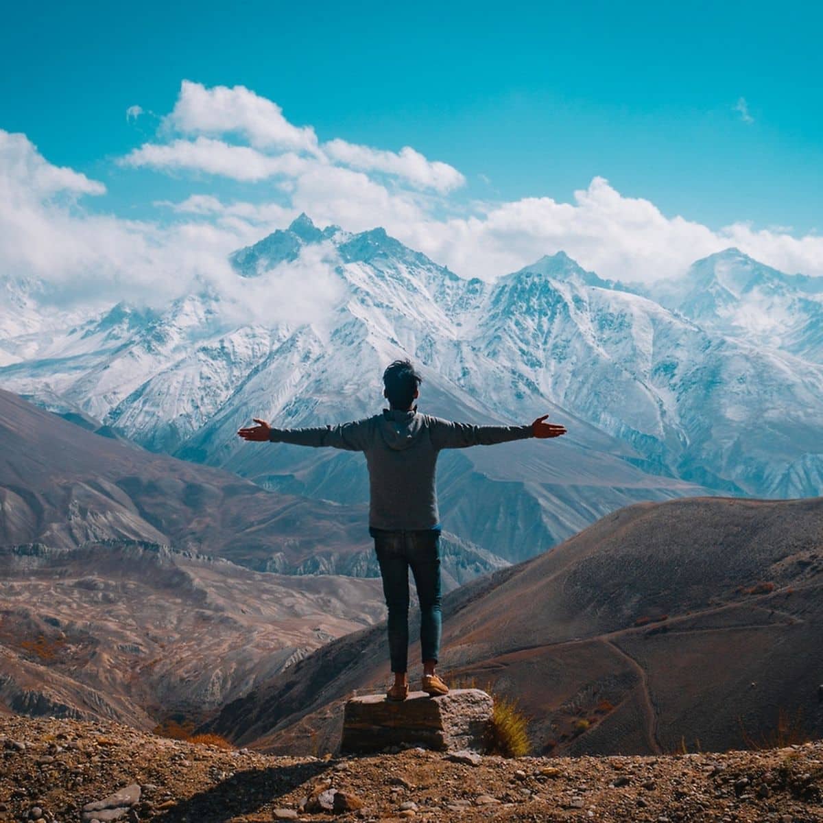 Person standing on a rock with arms outstretched, facing snow-capped mountains under a blue sky with scattered clouds.