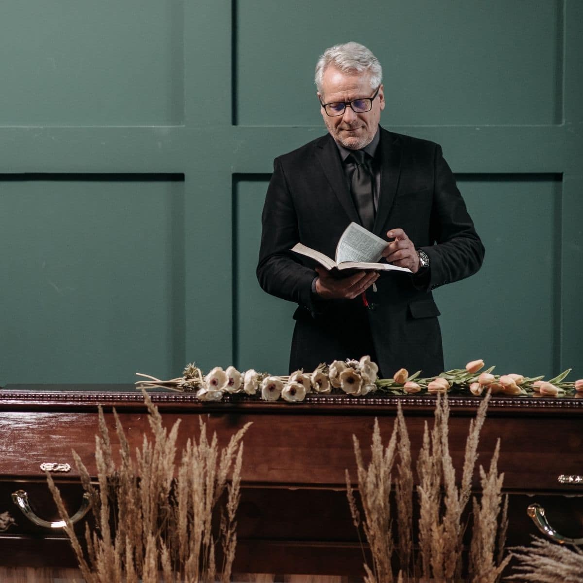 A man in a black suit stands behind a closed casket adorned with flowers, reading from a book during a funeral service.