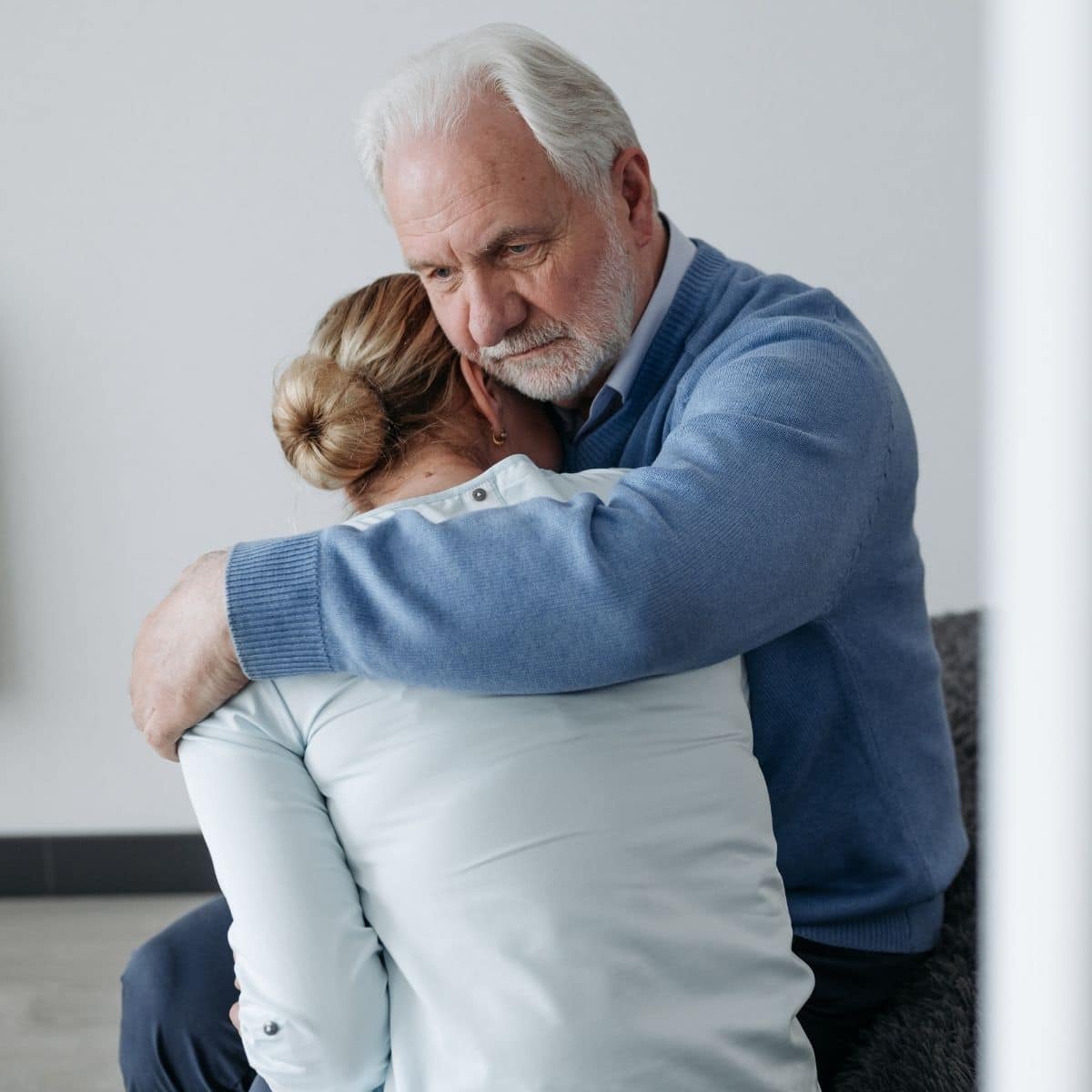 An elderly man with gray hair hugs a woman who is leaning her head on his chest. Both appear to be sitting and sharing a moment of comfort in a neutral indoor setting.