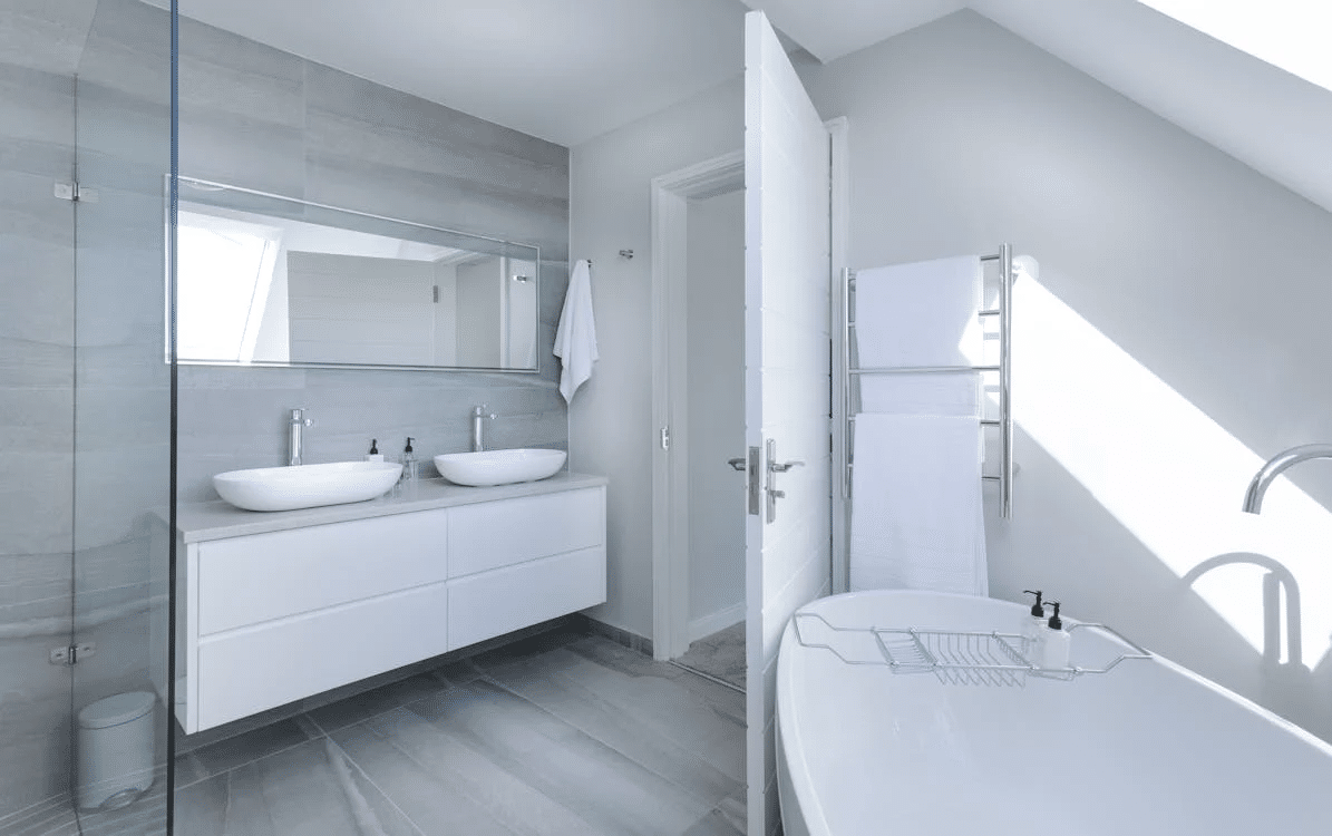 Modern bathroom with double sinks, a large mirror, white vanity, freestanding bathtub, towel rack with towels, gray tile floor, and natural light from a window.