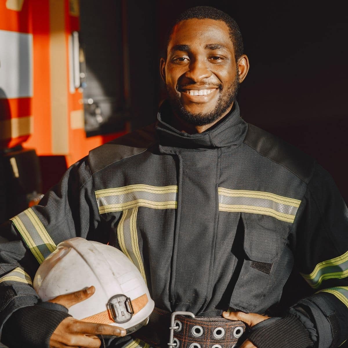 A firefighter in uniform stands indoors, holding a white helmet and smiling at the camera.