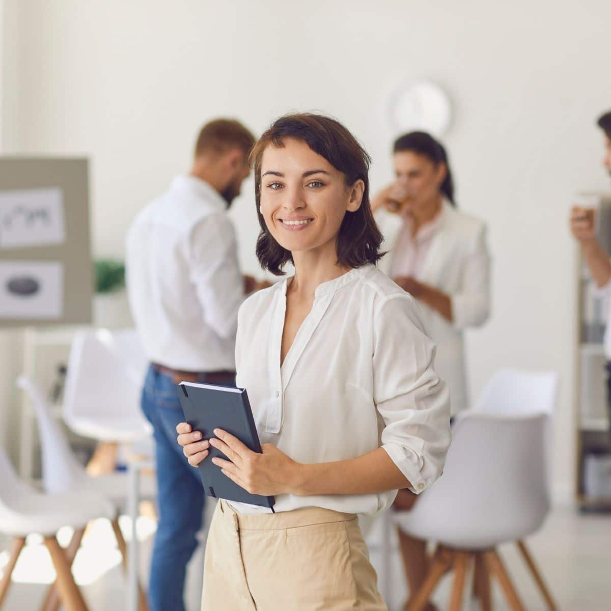 A woman stands in an office holding a tablet and smiling, while three colleagues talk and drink coffee in the background.