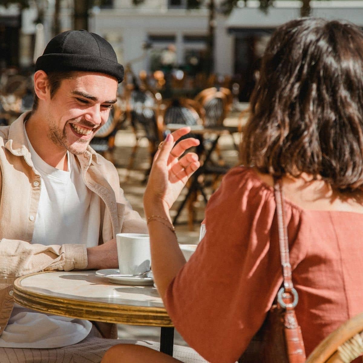 Two people sit at an outdoor café table, engaged in conversation and drinking from white cups. The man is smiling and wearing a black beanie; the woman has shoulder-length hair.