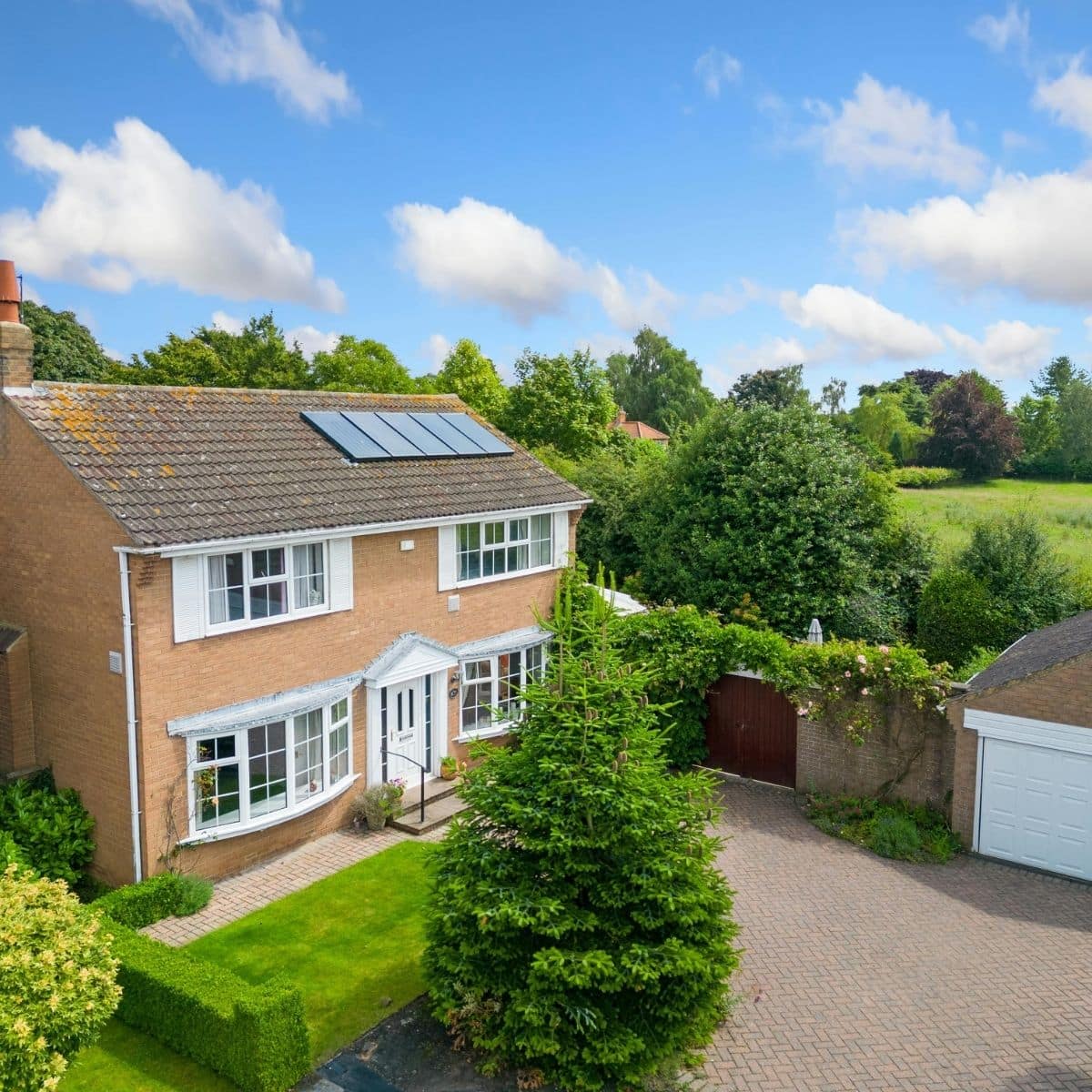 Two-story brick house with solar panels on the roof, a well-kept lawn, paved driveway, detached garage, and surrounding greenery under a blue sky with scattered clouds.