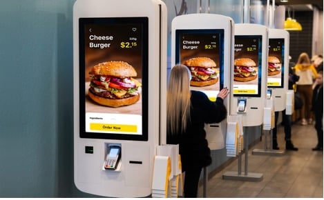A woman uses a digital self-service kiosk to order a cheeseburger in a fast food restaurant; several identical kiosks are visible in a row.