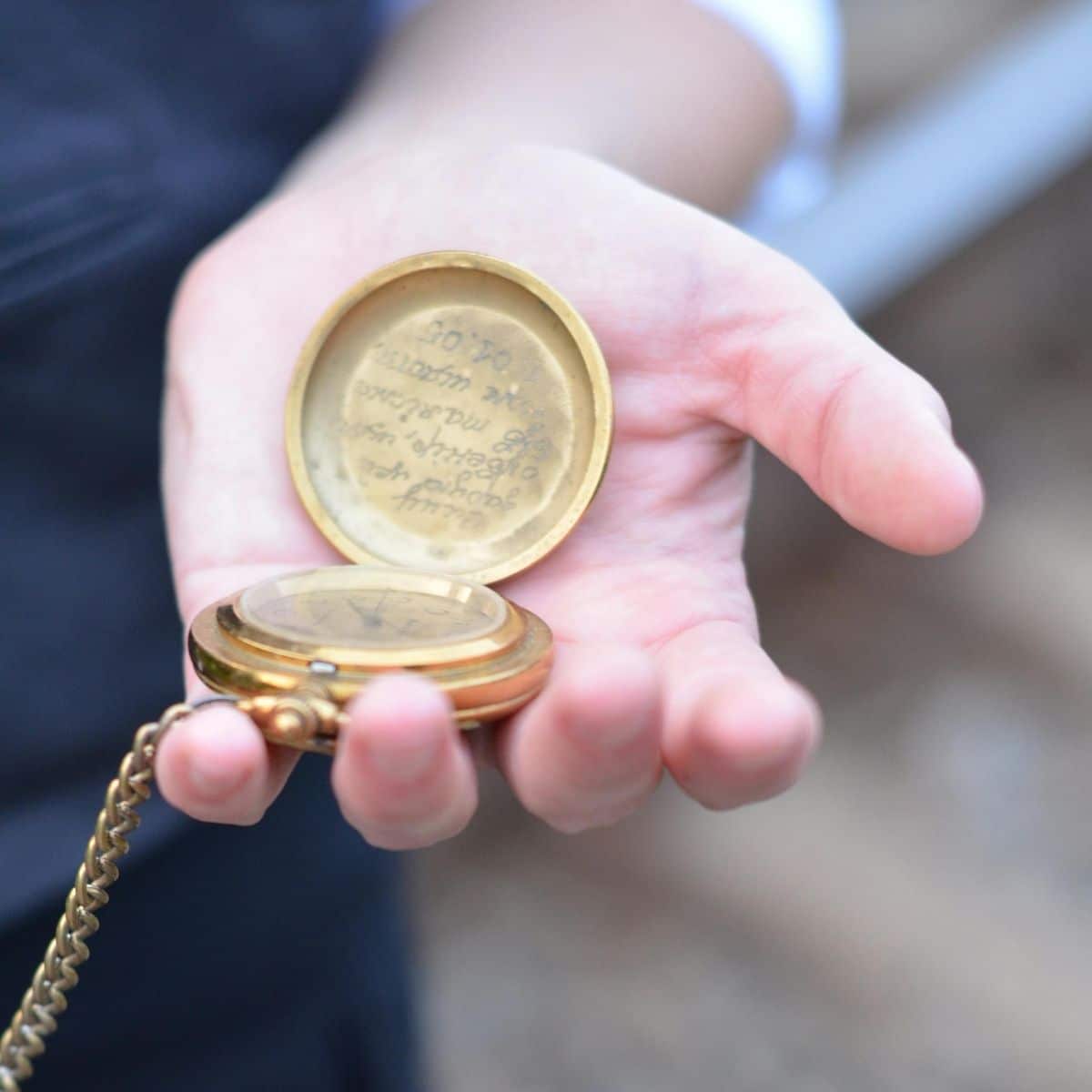 A hand holds an open gold pocket watch with an inscription on the inside cover and a visible chain attached.