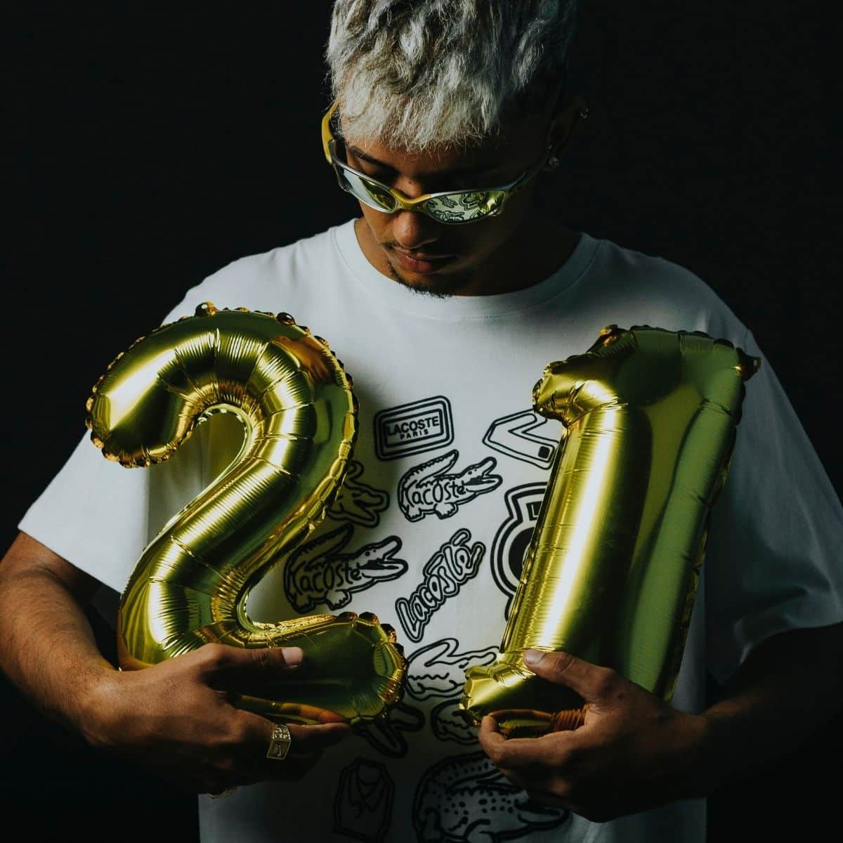 A person wearing sunglasses and a white graphic t-shirt holds gold foil balloons shaped like the numbers 2 and 1 against a dark background.