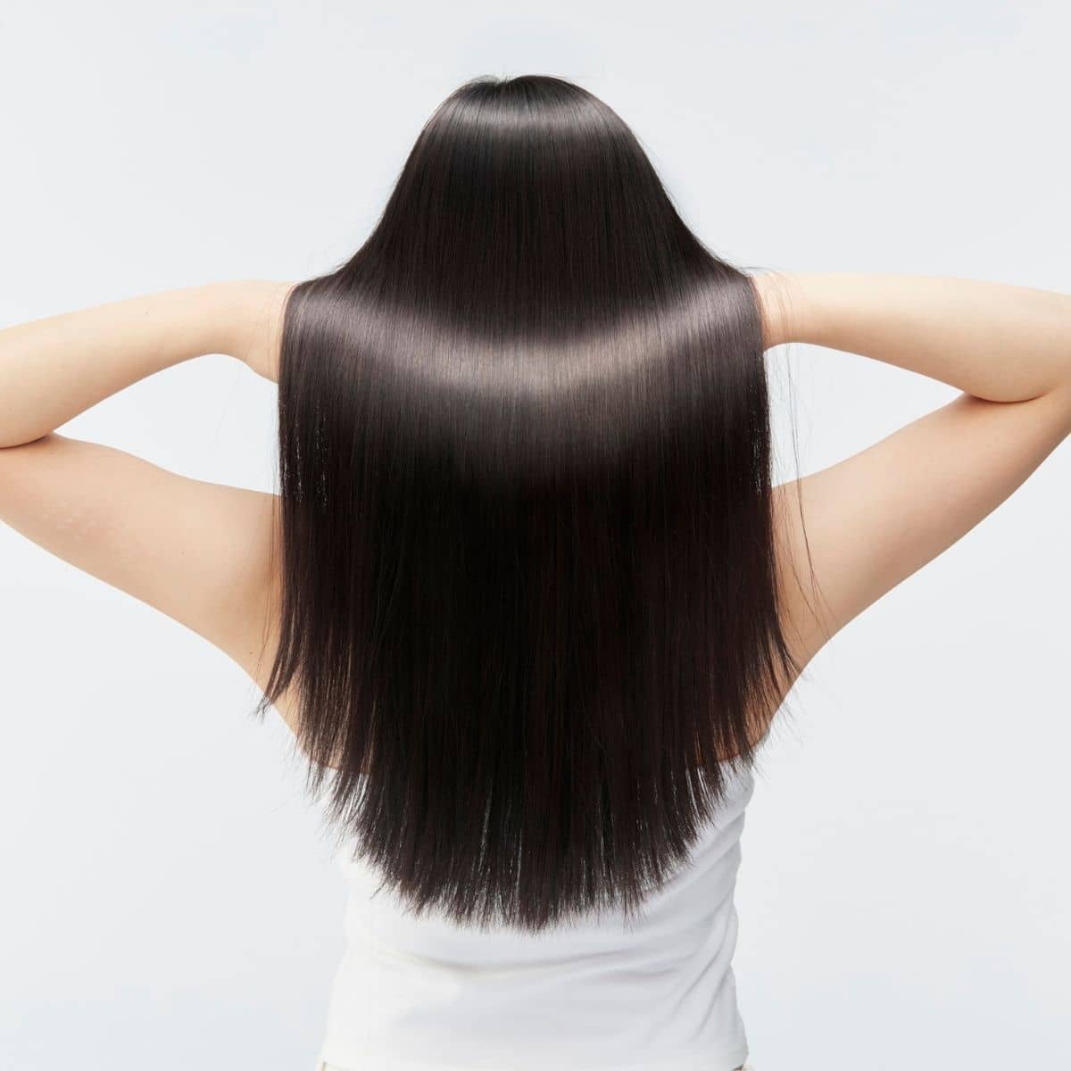 A person with long, straight, shiny black hair is seen from behind, holding their hair up with both hands and wearing a white sleeveless top.