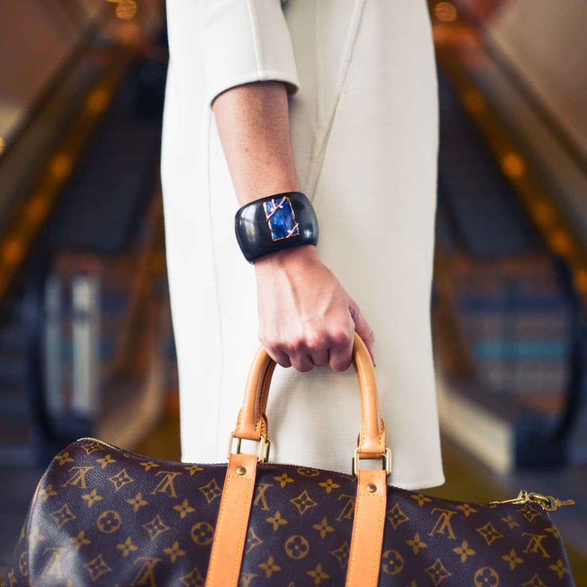 A person wearing a white coat holds a brown Louis Vuitton bag and wears a black smart wristband, standing in front of an escalator.