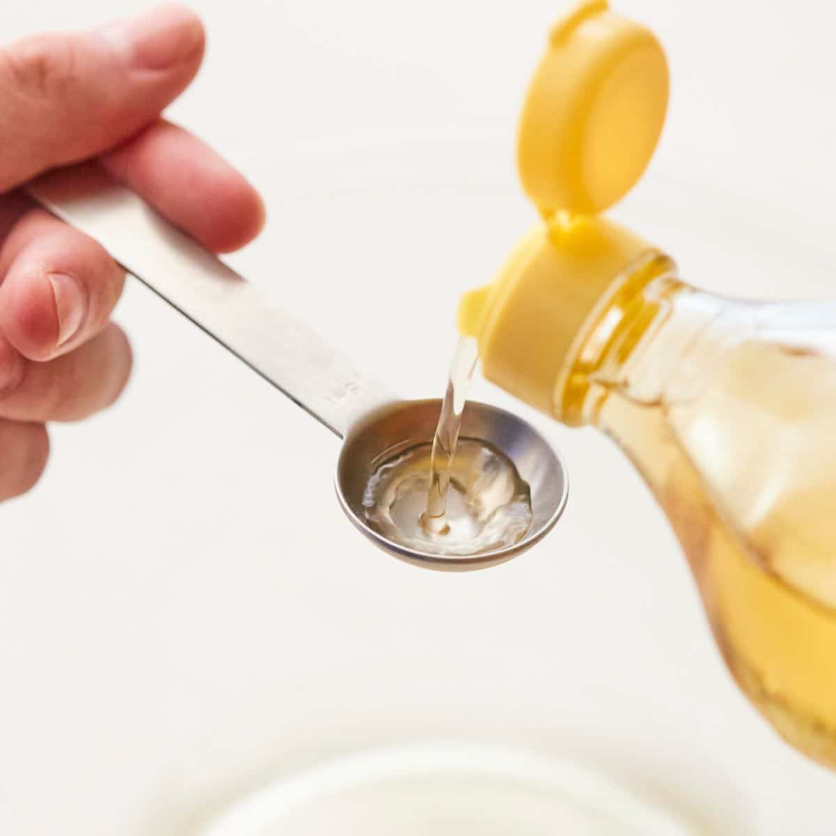 A hand pours a clear liquid from a yellow-capped bottle into a metal measuring spoon.