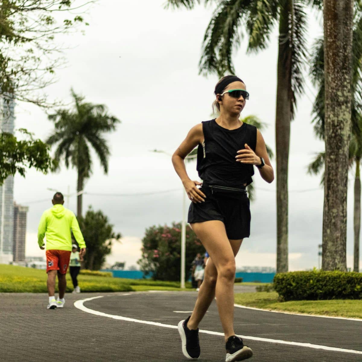 A woman in black athletic wear and sunglasses jogs on a paved path lined with palm trees; another person walks in the background.
