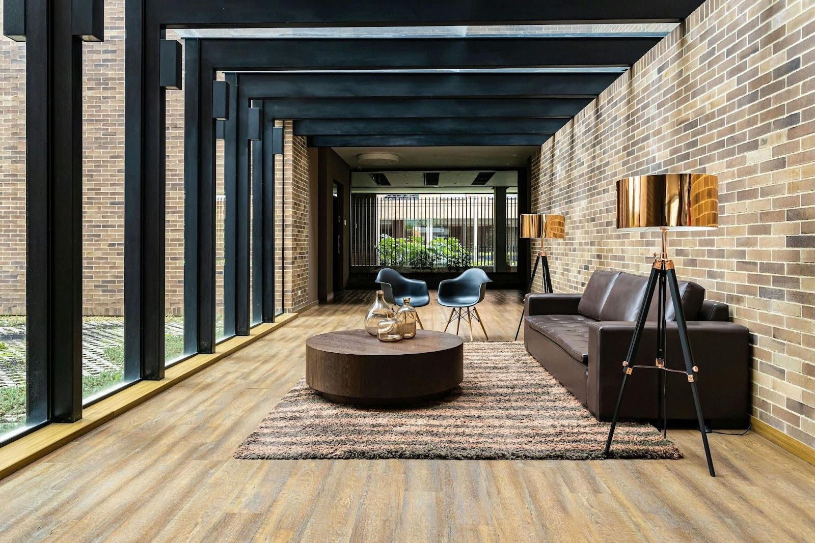 Modern lounge area with brown leather sofa, round ottoman, shaggy rug, two black chairs, and two gold floor lamps, set against brick walls and large windows with black frames.