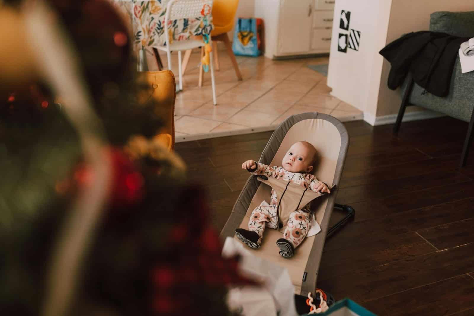 A baby in a patterned onesie sits in a baby bouncer on a wooden floor, looking toward a decorated Christmas tree in a living room.