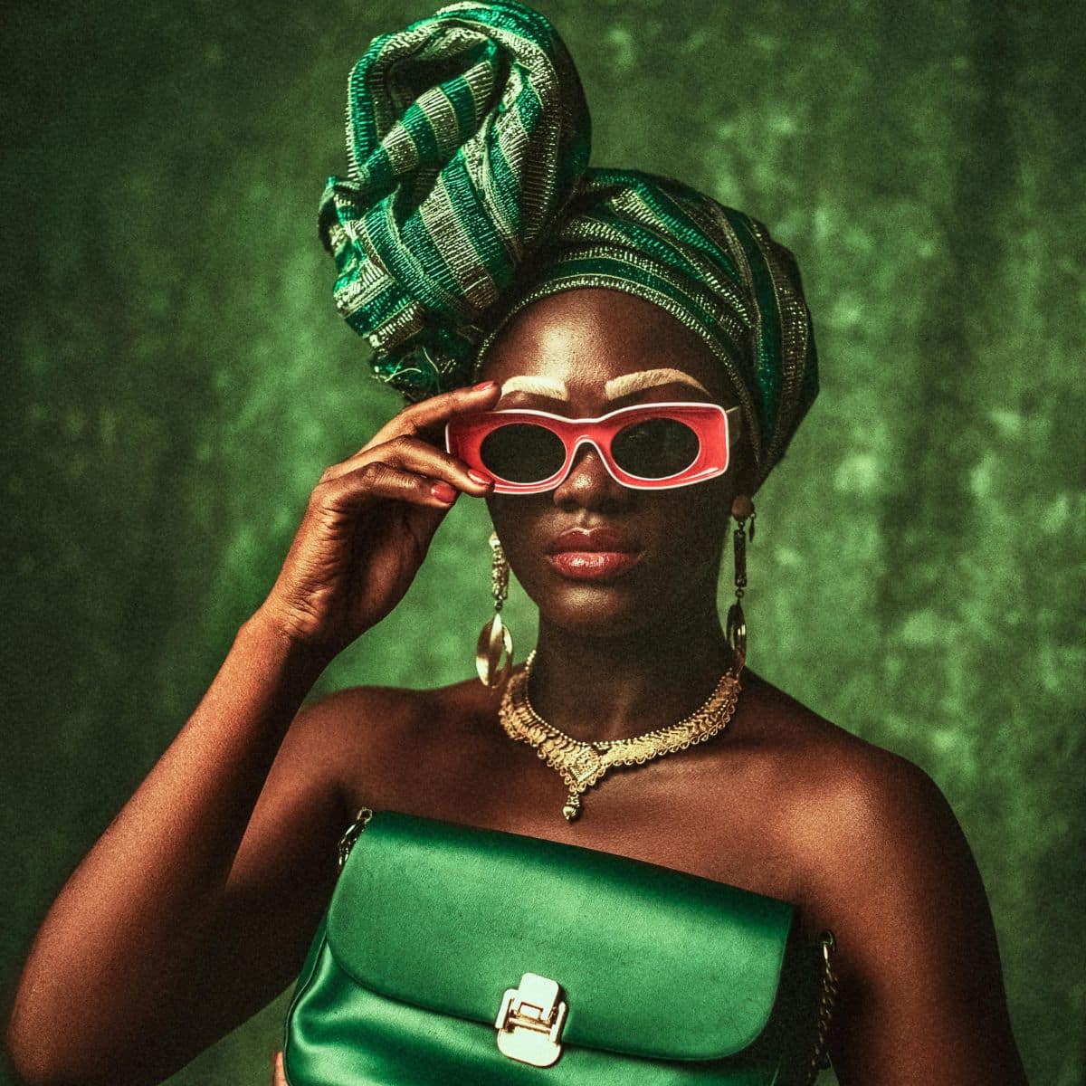 A woman wearing a green headwrap and holding a green purse poses against a green background. She wears red sunglasses, gold jewelry, and has bold white eyebrows.