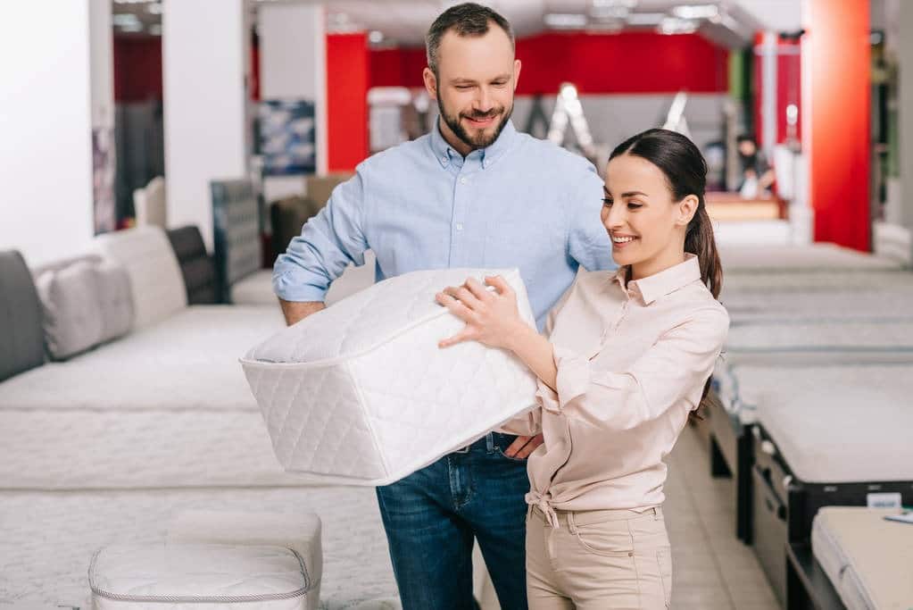A man and woman in a mattress store examine a white mattress sample together, surrounded by display beds in the showroom.