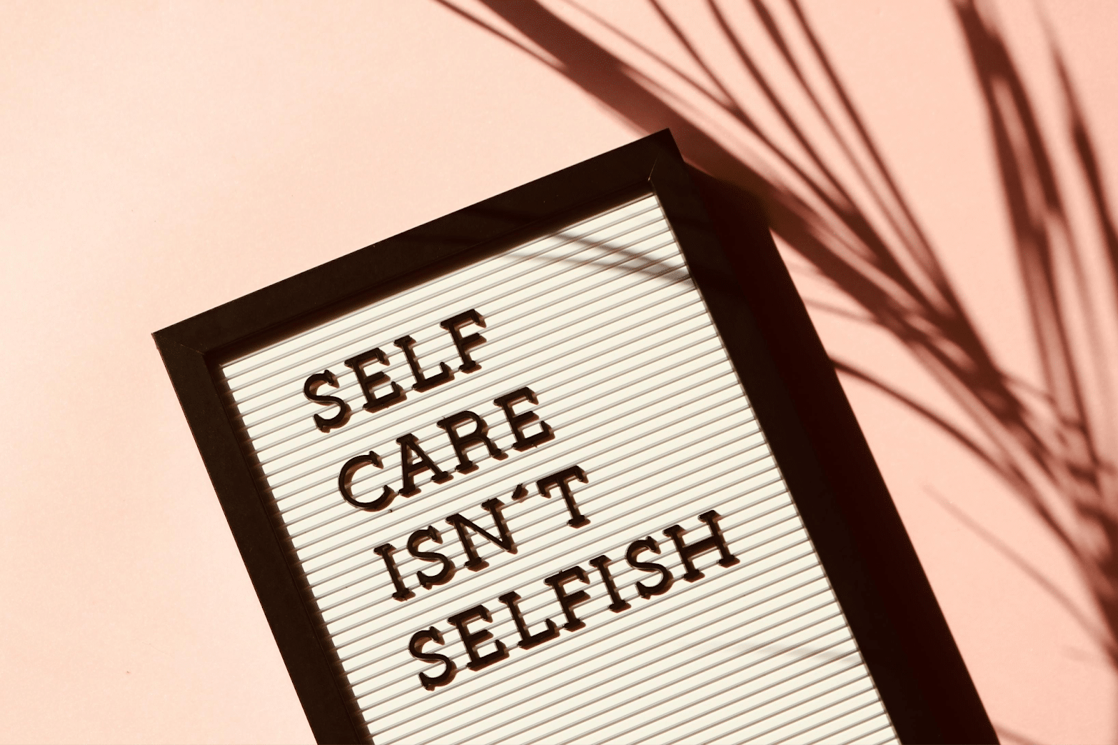 A letter board on a pink background displays the message "SELF CARE ISN'T SELFISH" in black capital letters, with a shadow of a plant crossing the scene.