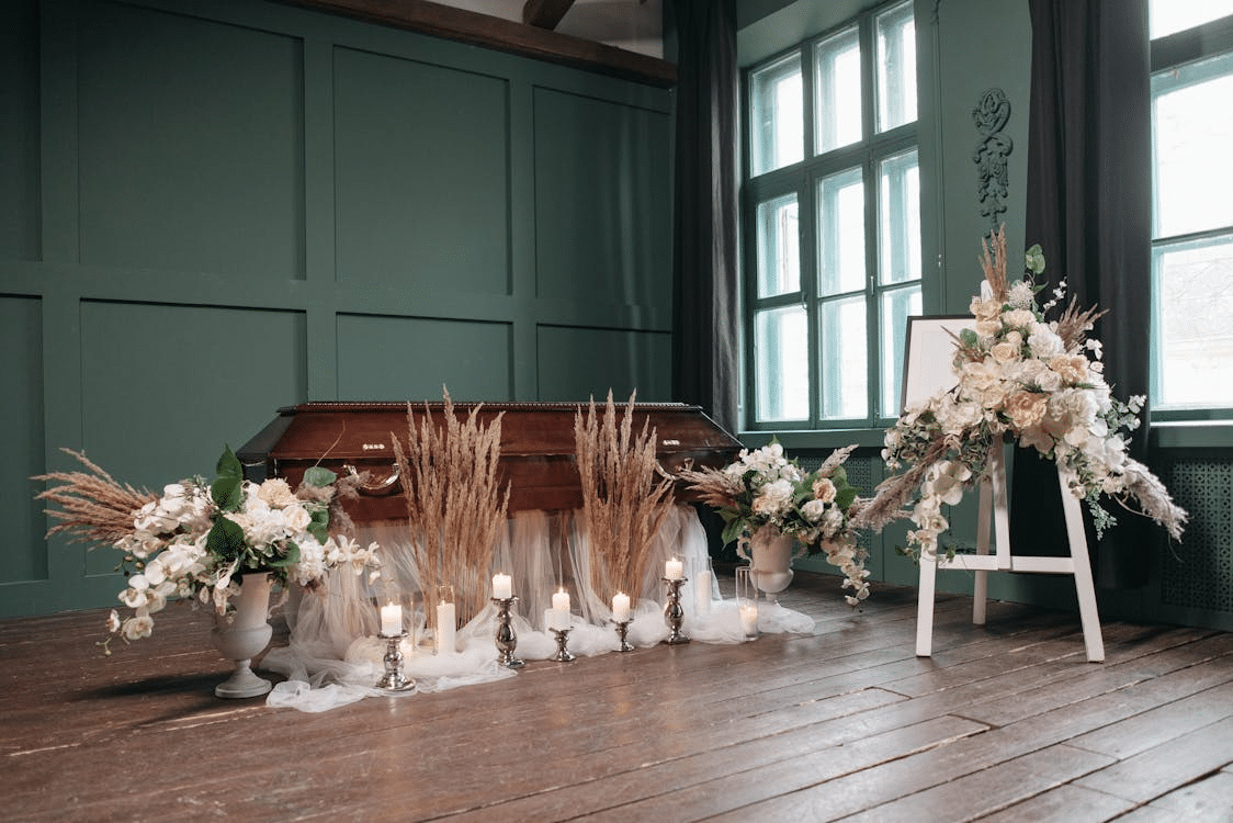 A wooden coffin is surrounded by white floral arrangements, candles, and an easel with flowers in a softly lit room with wooden floors and green walls.