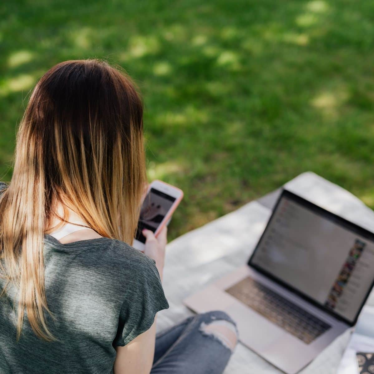 Person sitting outdoors on a blanket, using a smartphone in one hand and a laptop in front of them, with grass in the background.