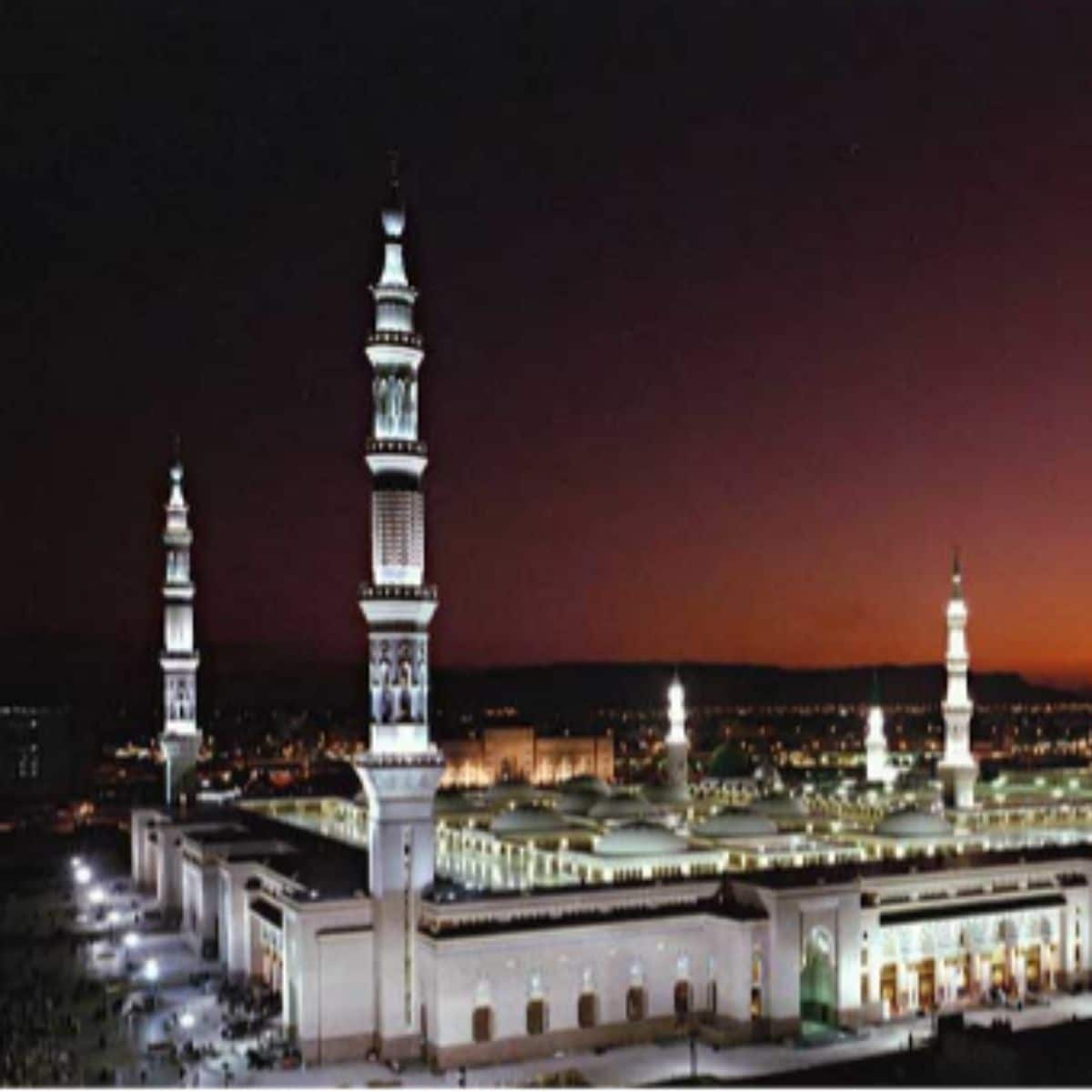 A nighttime view of Al-Masjid an-Nabawi in Medina, featuring illuminated minarets and mosque buildings against a dark sky.