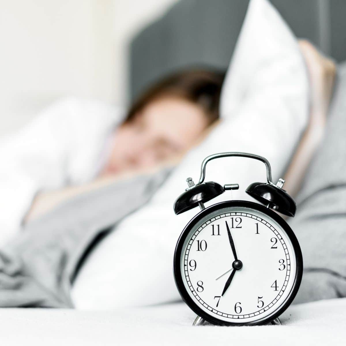 A black alarm clock shows 7:00 while a person lies in bed under a grey blanket, partially blurred in the background.