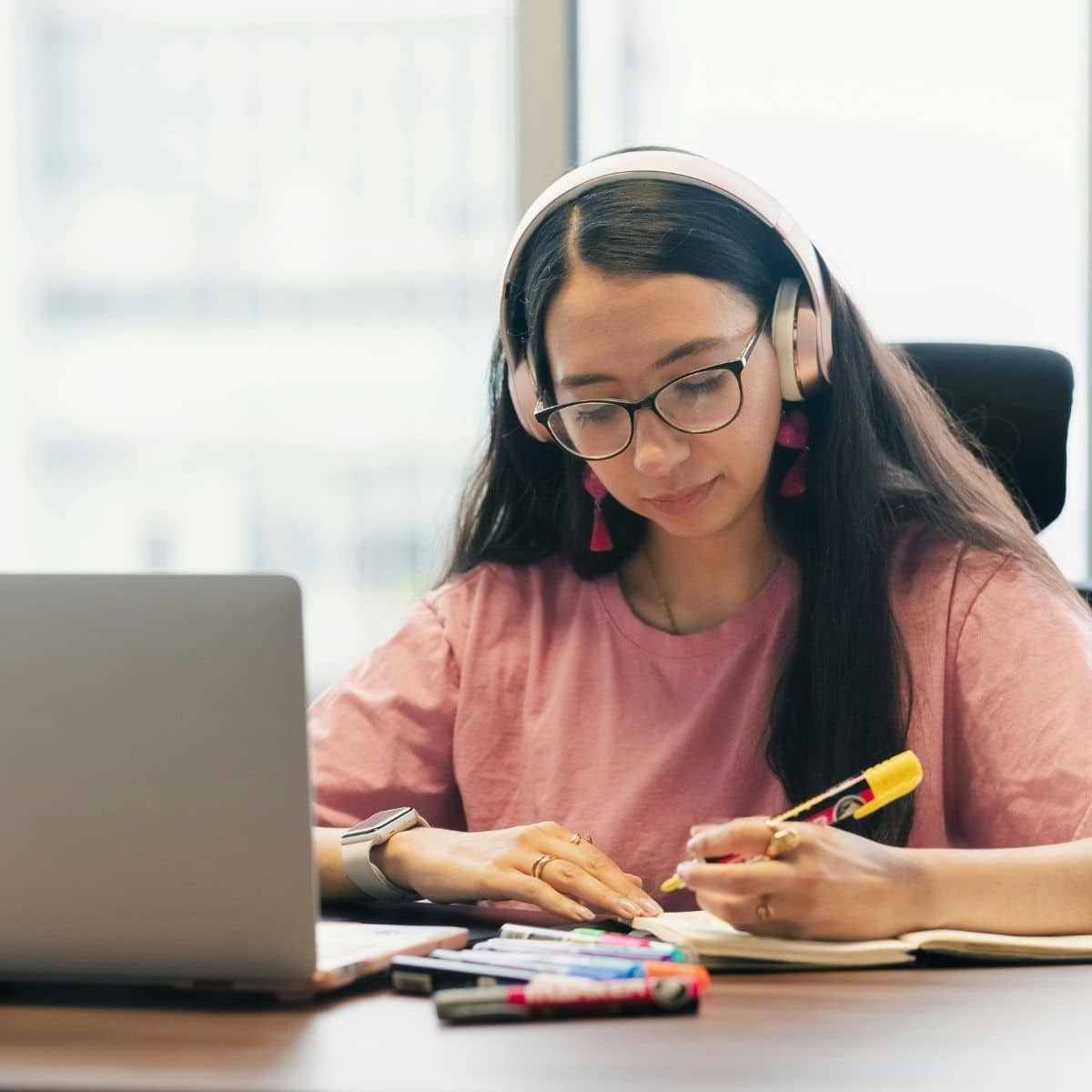 A person wearing headphones and a pink shirt writes in a notebook at a desk with a laptop and markers.