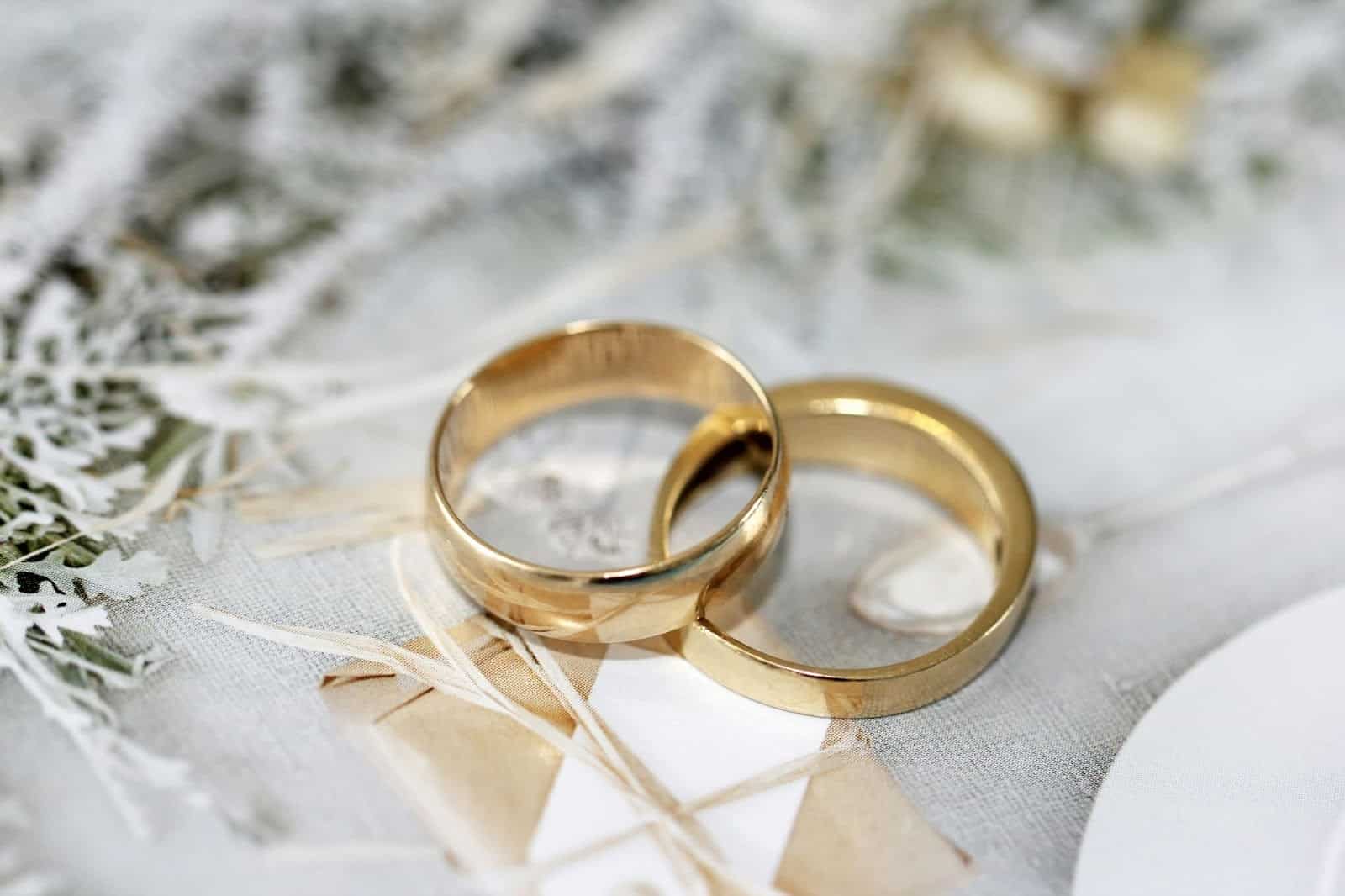 Two gold wedding bands are placed next to each other on a light-colored, textured surface with decorative elements in the background.