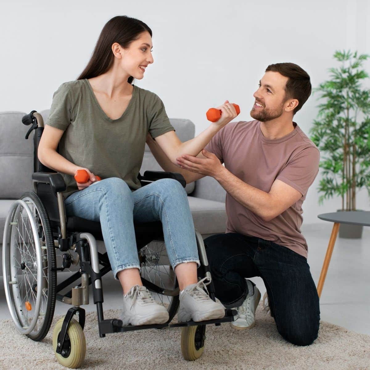 A woman in a wheelchair lifts a small dumbbell while a man kneels beside her, assisting with her exercise in a sitting room.