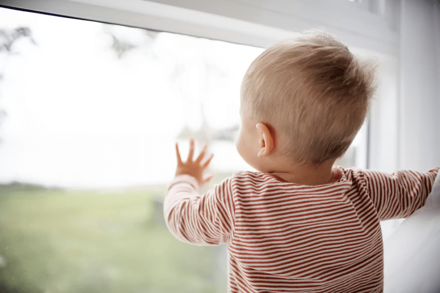 A young child in a striped shirt stands indoors, facing and touching a window whilst looking outside.