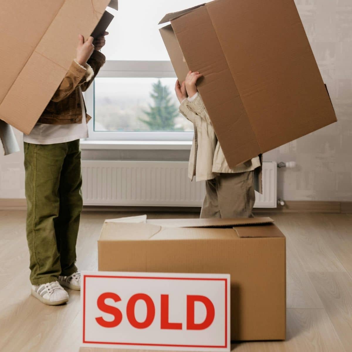 Two children stand indoors with cardboard boxes, one box covering a child's head, next to a large "SOLD" sign, suggesting a recent house purchase.