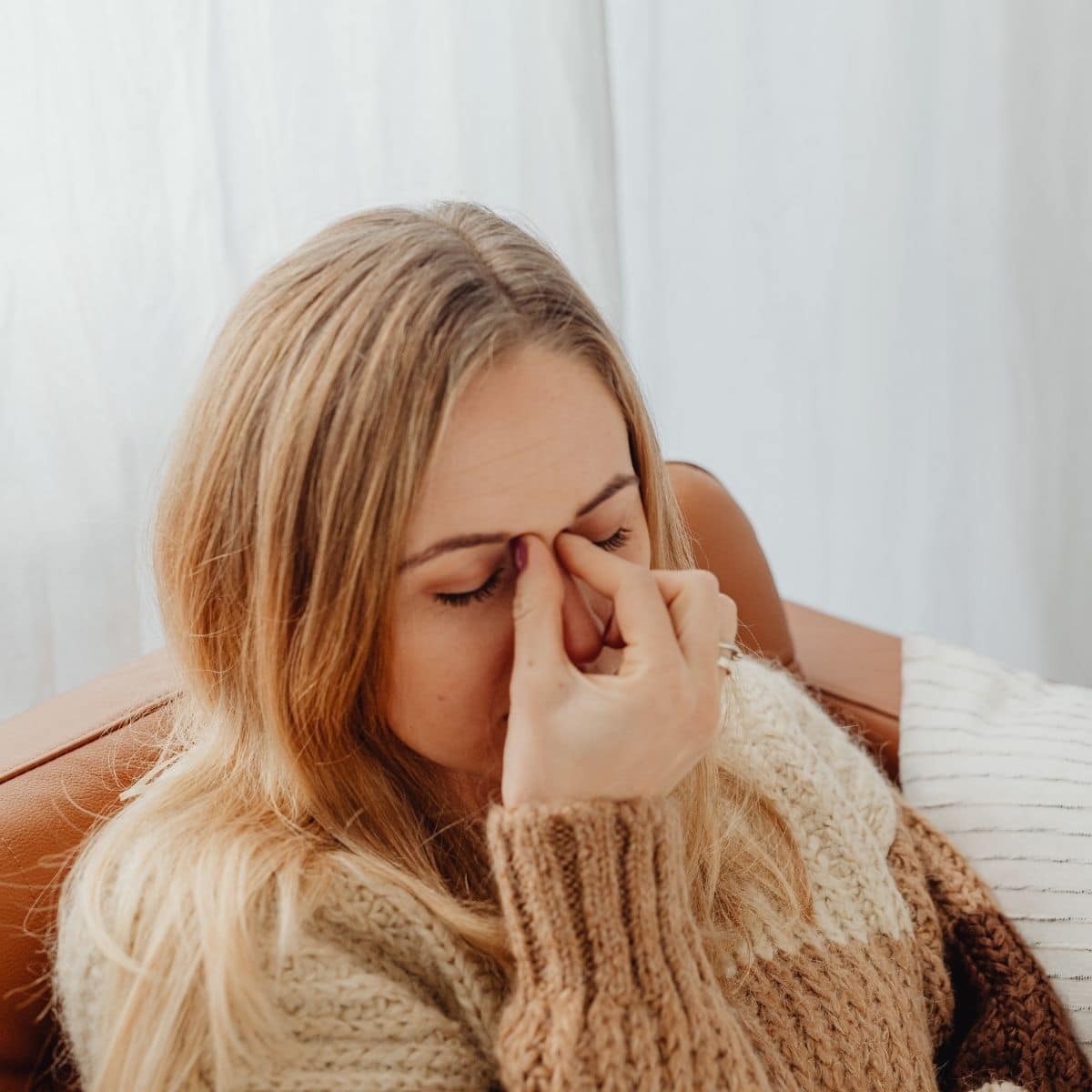 A woman sits on a brown sofa, holding the bridge of her nose with her eyes closed, appearing to be stressed or fatigued.