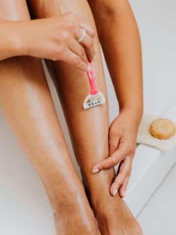 A person shaves their lower leg with a pink razor, using their other hand to stretch the skin. A round soap rests nearby on a flannel.