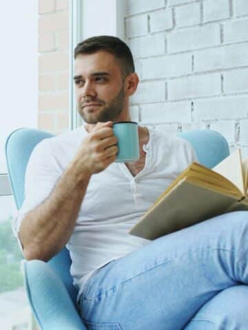 Man sitting in a light blue armchair by a window, holding a mug and an open book, looking to the side.