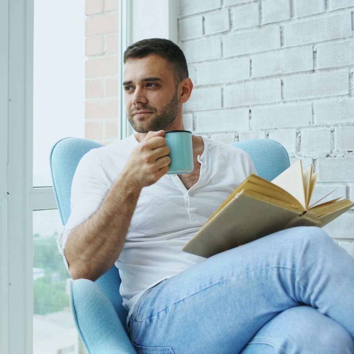 Man sitting in a light blue armchair by a window, holding a mug and an open book, looking to the side.